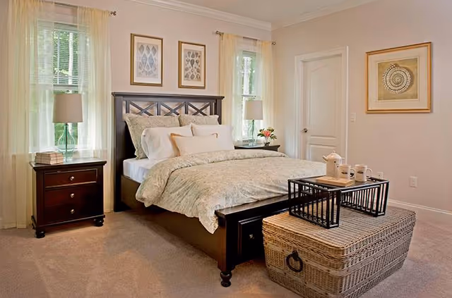 A cozy bedroom with a dark wooden bed frame and a neatly made bed with light-colored bedding. There are two windows with sheer curtains on either side of the bed, a nightstand with a lamp and books on the left, and a wicker chest at the foot of the bed with a black tray holding a teapot and cups. The walls are light-colored with framed artwork hanging above the bed and on the adjacent wall.