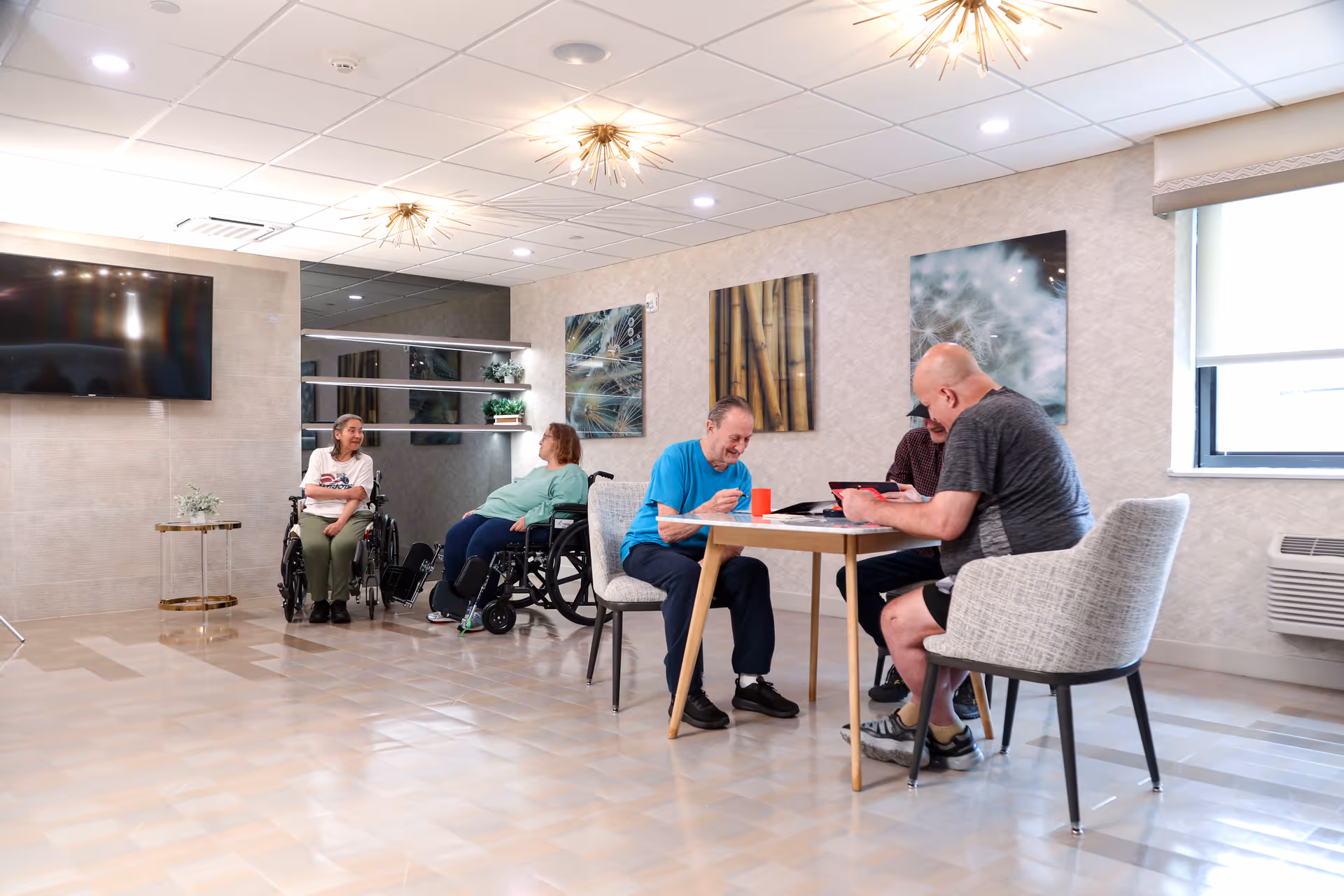 A bright common area in a senior living facility with five elderly individuals, two in wheelchairs and three seated around a table engaged in activities. The room has modern decor with abstract wall art, a large flat-screen TV mounted on the wall, and stylish light fixtures on the ceiling.