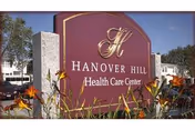Maroon entrance sign reading "Hanover Hill Health Care Center" with flowers in front and buildings and blue sky behind.
