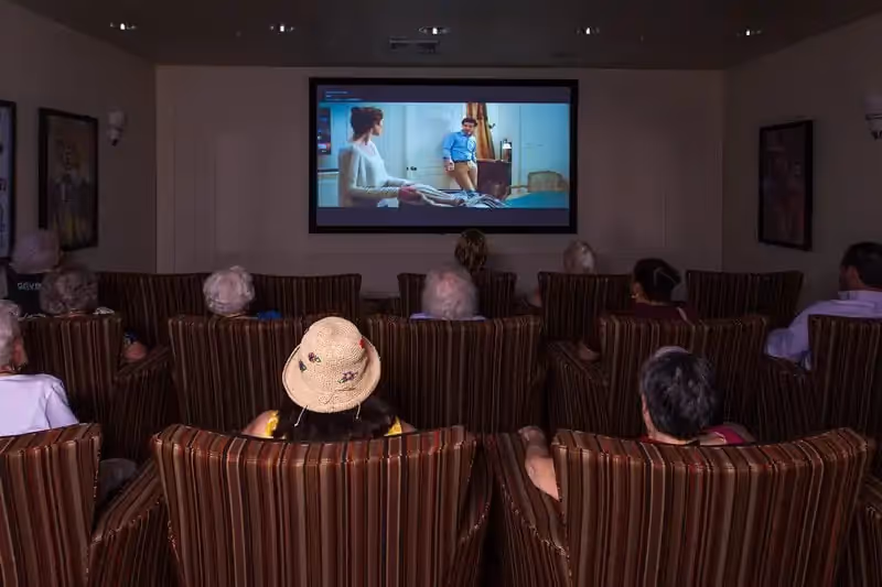 A group of elderly people seated in striped armchairs watching a movie projected on a large screen in a dimly lit room.