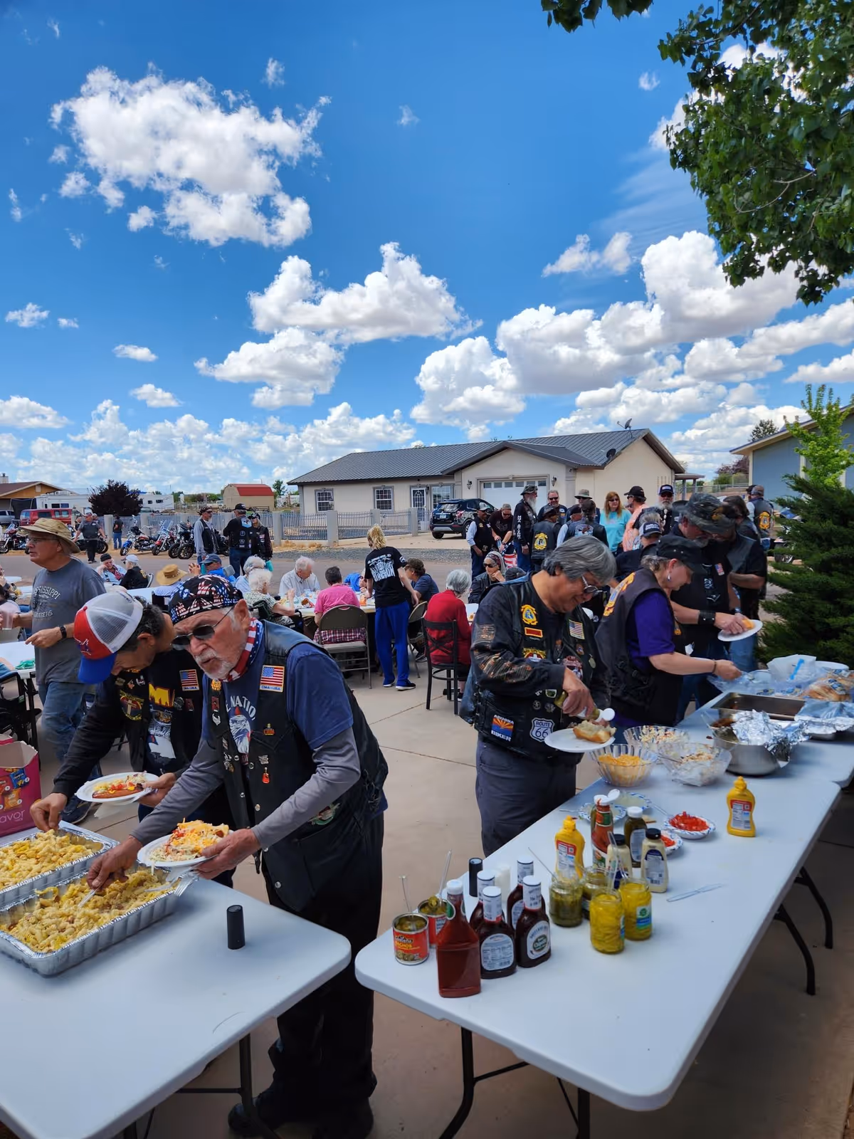 A group of people gathered outdoors at Carriage House On West Garden Lane, serving themselves food from buffet tables set up with various condiments and dishes. The sky is bright blue with scattered clouds, and there are houses and vehicles in the background. Many attendees are wearing biker vests and hats.