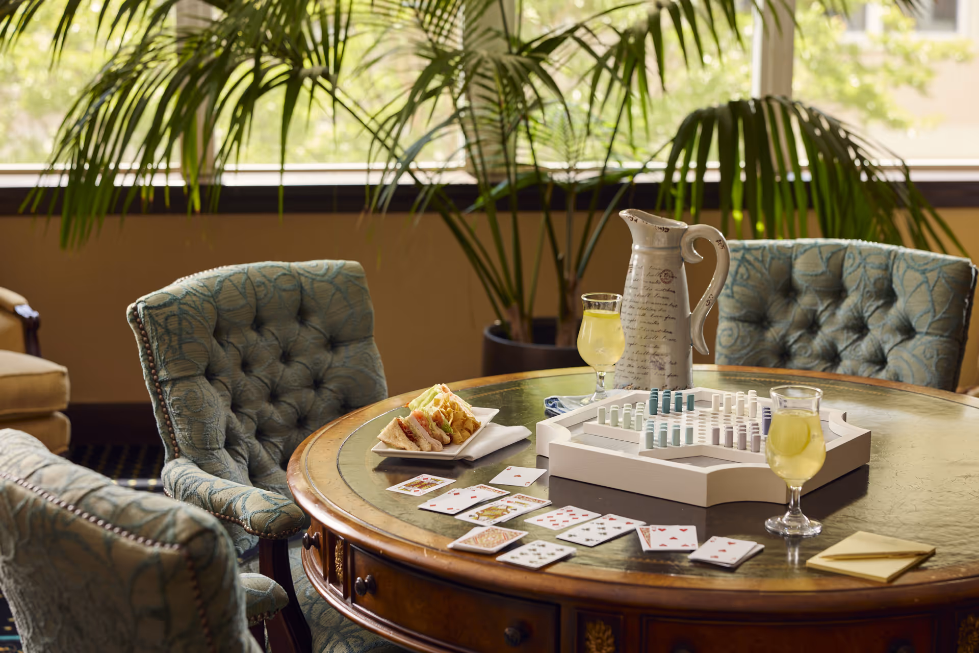 A round wooden table with a green leather inset, surrounded by upholstered chairs with a blue patterned fabric. On the table are two glasses of a light yellow beverage, a decorative pitcher, a plate with sandwiches and chips, a board game with pieces set up, and playing cards spread out. In the background, large green plants and a window letting in natural light are visible.