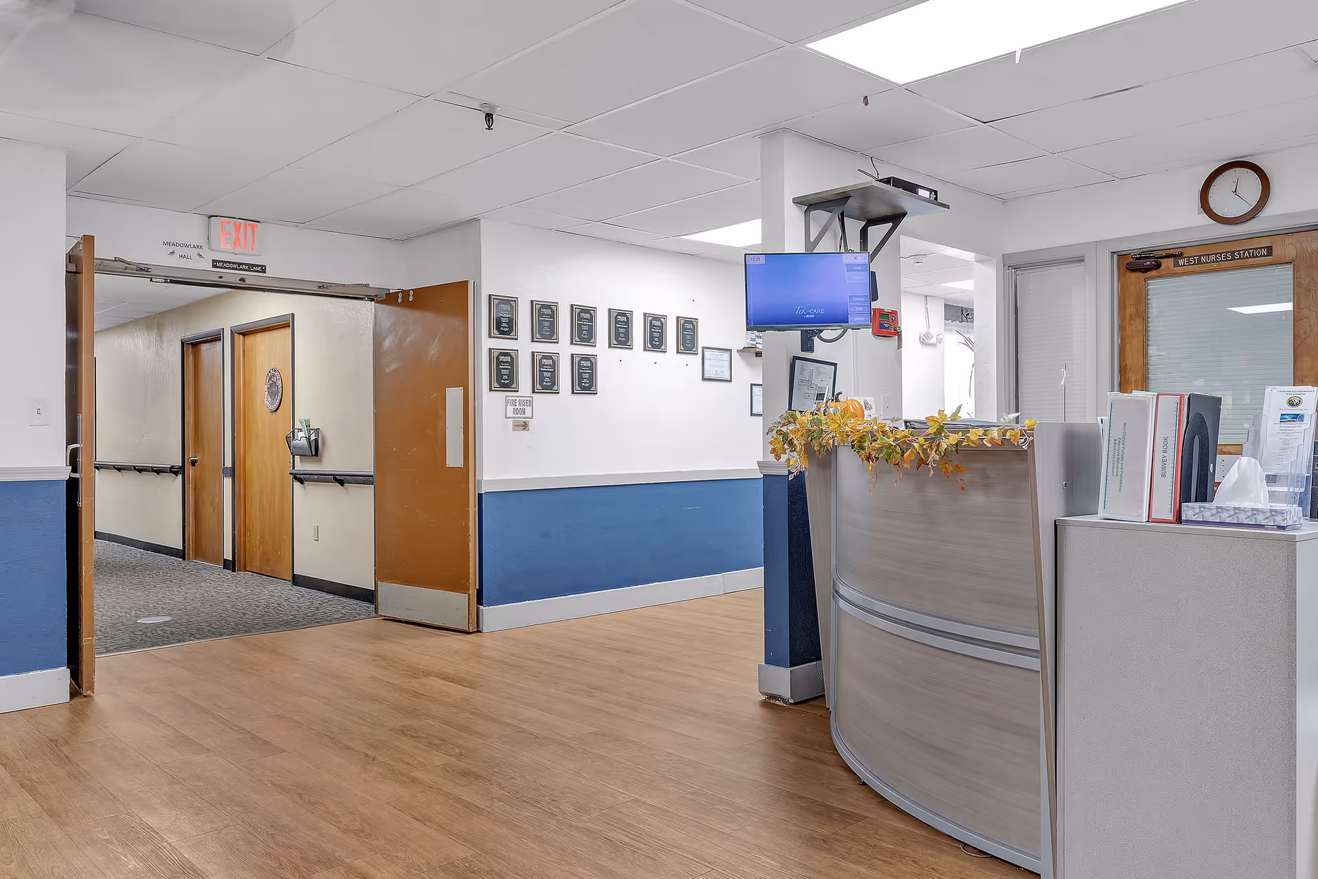 Interior view of a senior care facility hallway with a nurse's station on the right. The nurse's station has a curved desk with office supplies and a monitor mounted above it. There are plaques on the wall and an exit sign above double doors leading to another hallway with wooden doors.