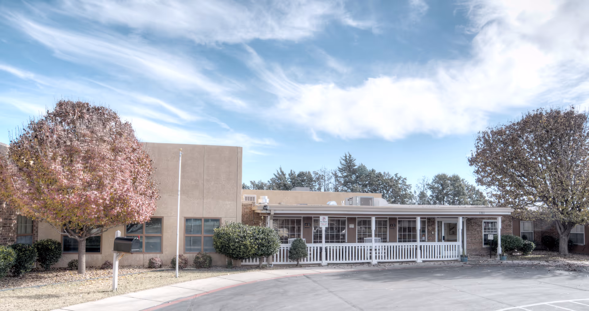 Single-story healthcare facility with a white-railed front porch, trees on either side, and a paved driveway under a blue sky.