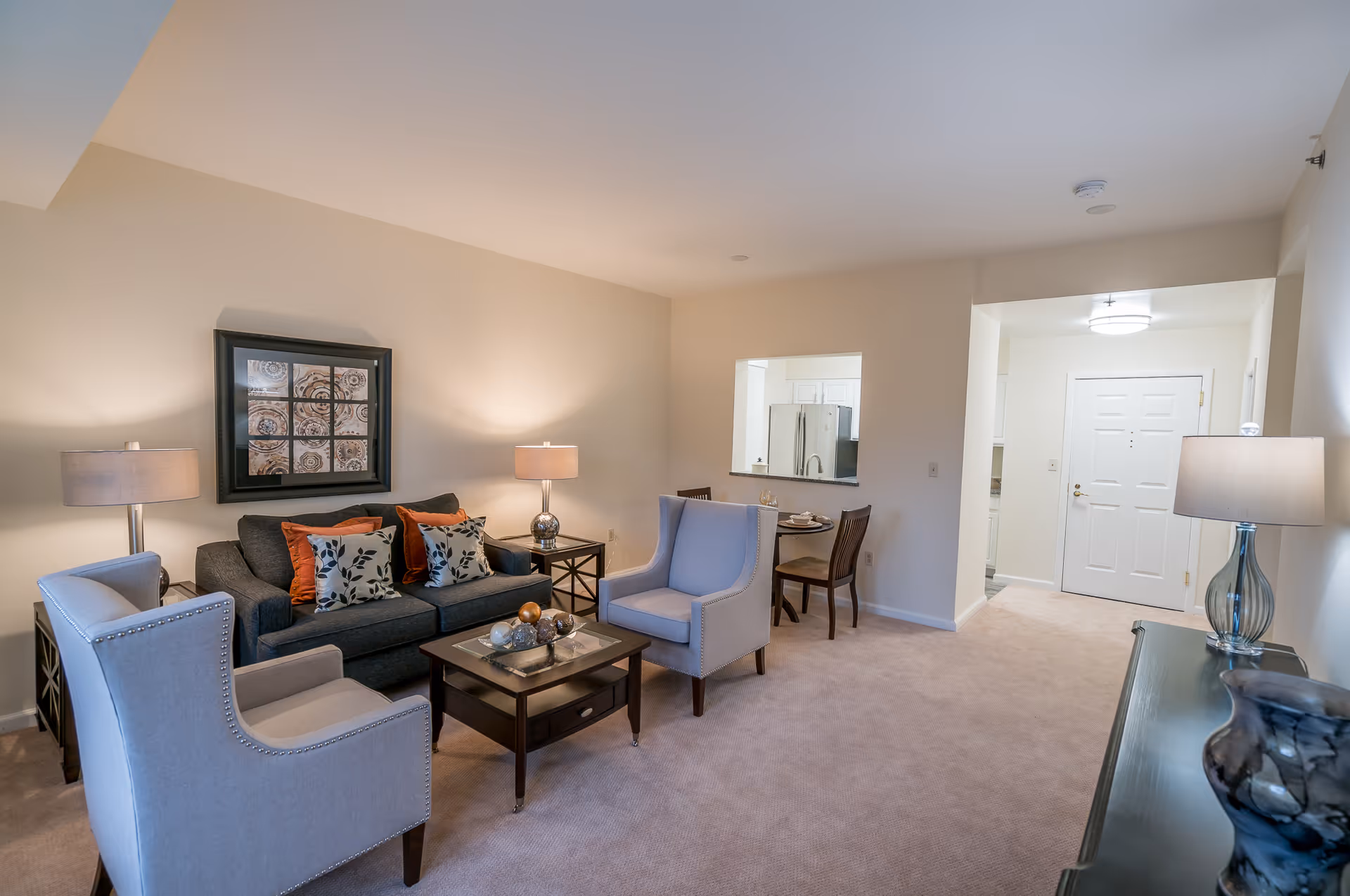 Well-lit furnished living room with a sofa, armchairs, coffee table, lamps, and a view toward the entry and kitchen pass-through.