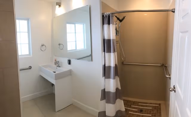 A clean bathroom with a white sink and a large mirror above it. There is a window next to the sink allowing natural light in. To the right, there is a walk-in shower with a striped shower curtain and grab bars installed on the walls for safety.