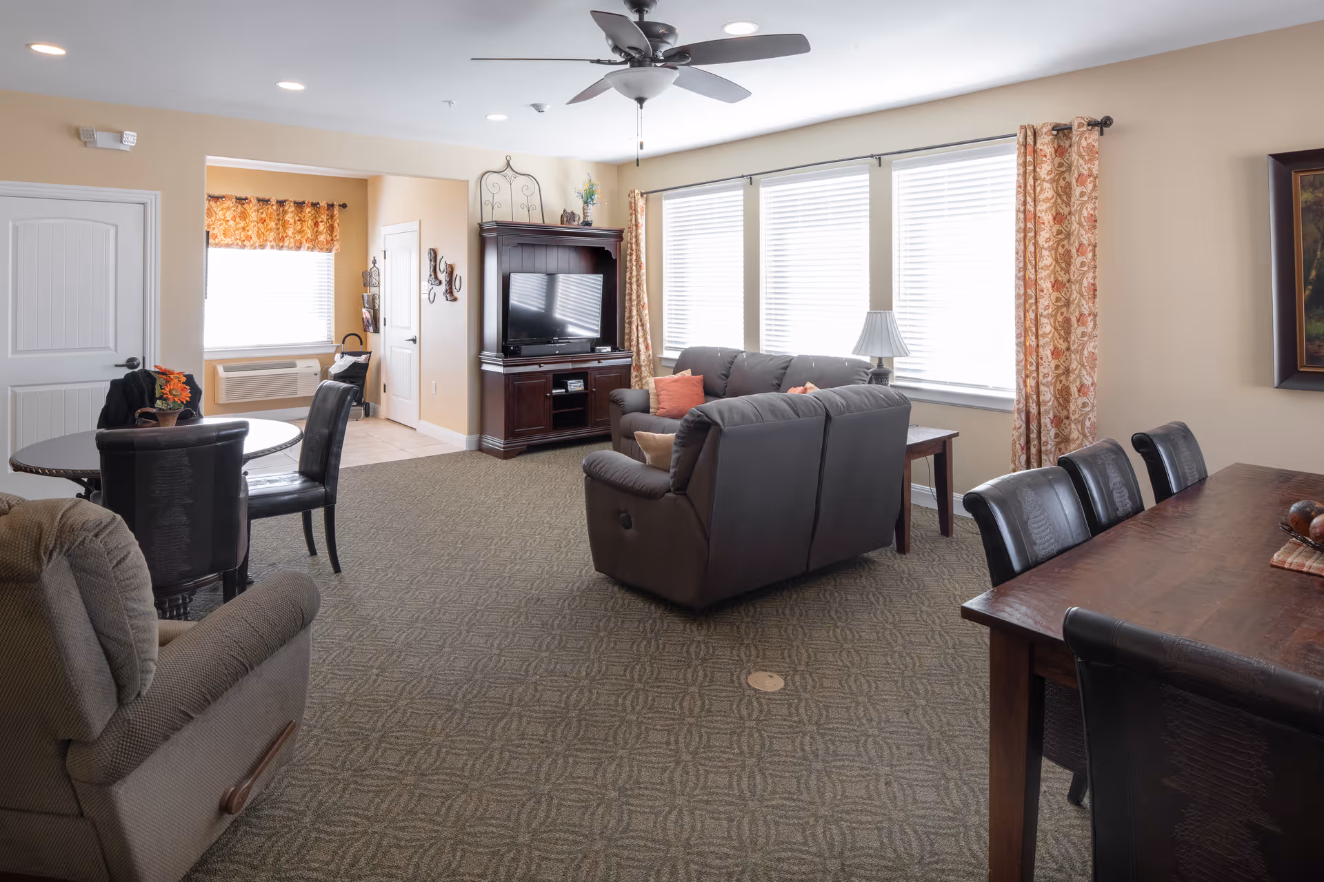 A spacious living room area in an assisted living facility with a ceiling fan, large windows with blinds and patterned curtains, a dark brown leather sofa with cushions, a wooden TV cabinet with a flat-screen TV, a round table with chairs, and a dining table with chairs. The room has beige walls and carpeted flooring.