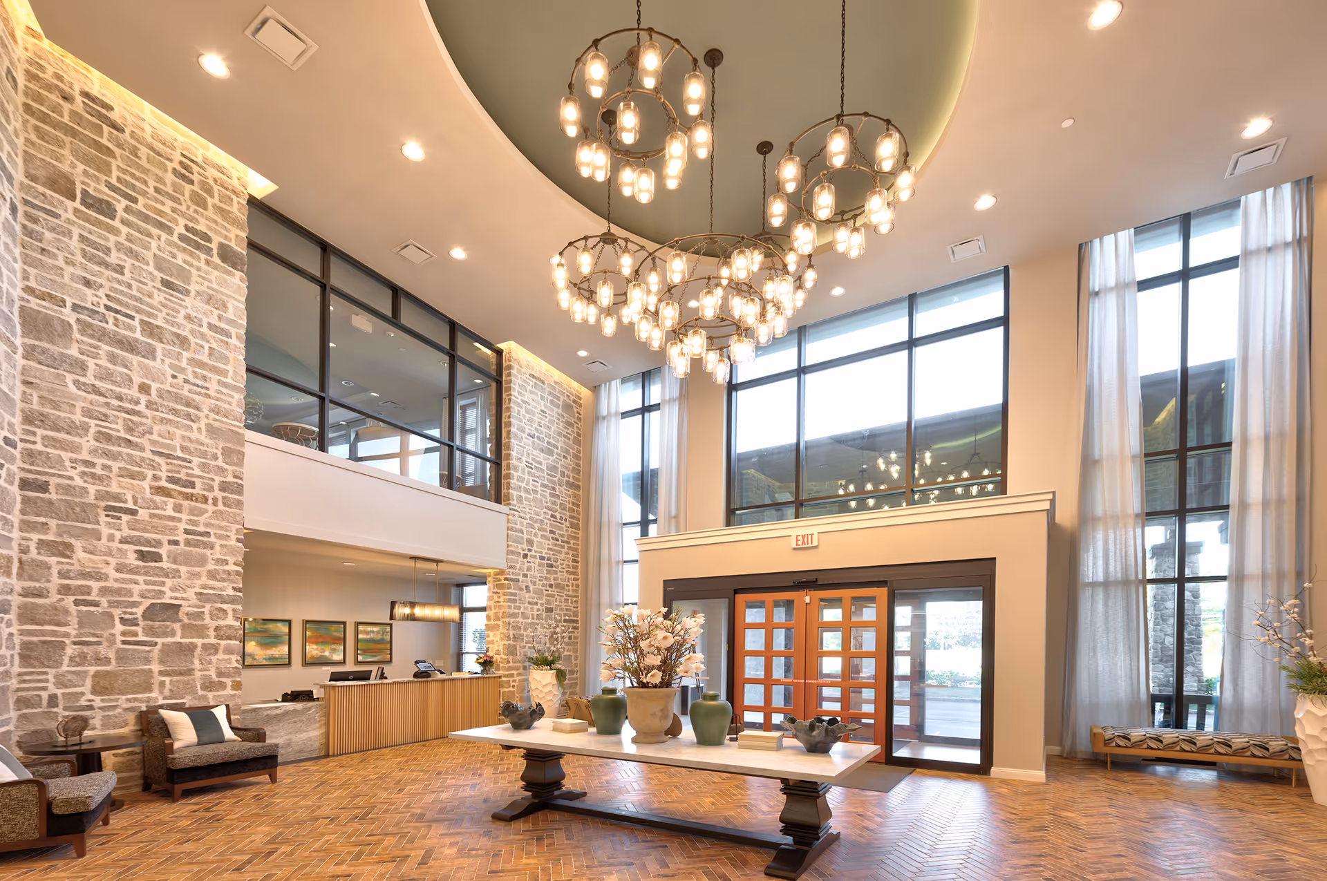 Spacious and elegant lobby area of a senior living facility with high ceilings, large windows, stone accent walls, a central table decorated with vases and flowers, comfortable seating, and modern chandeliers hanging from the ceiling.