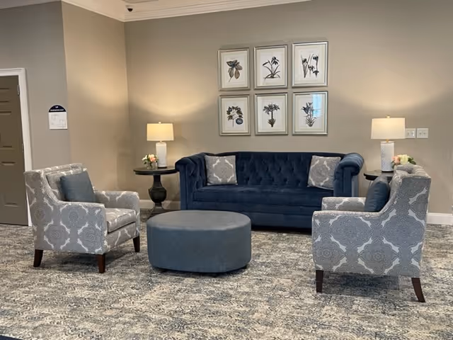 A cozy seating area with a dark blue tufted sofa, two patterned armchairs with blue cushions, and a round blue ottoman in the center. There are two side tables with lamps and small flower arrangements. Six framed botanical prints hang on the beige wall behind the sofa.