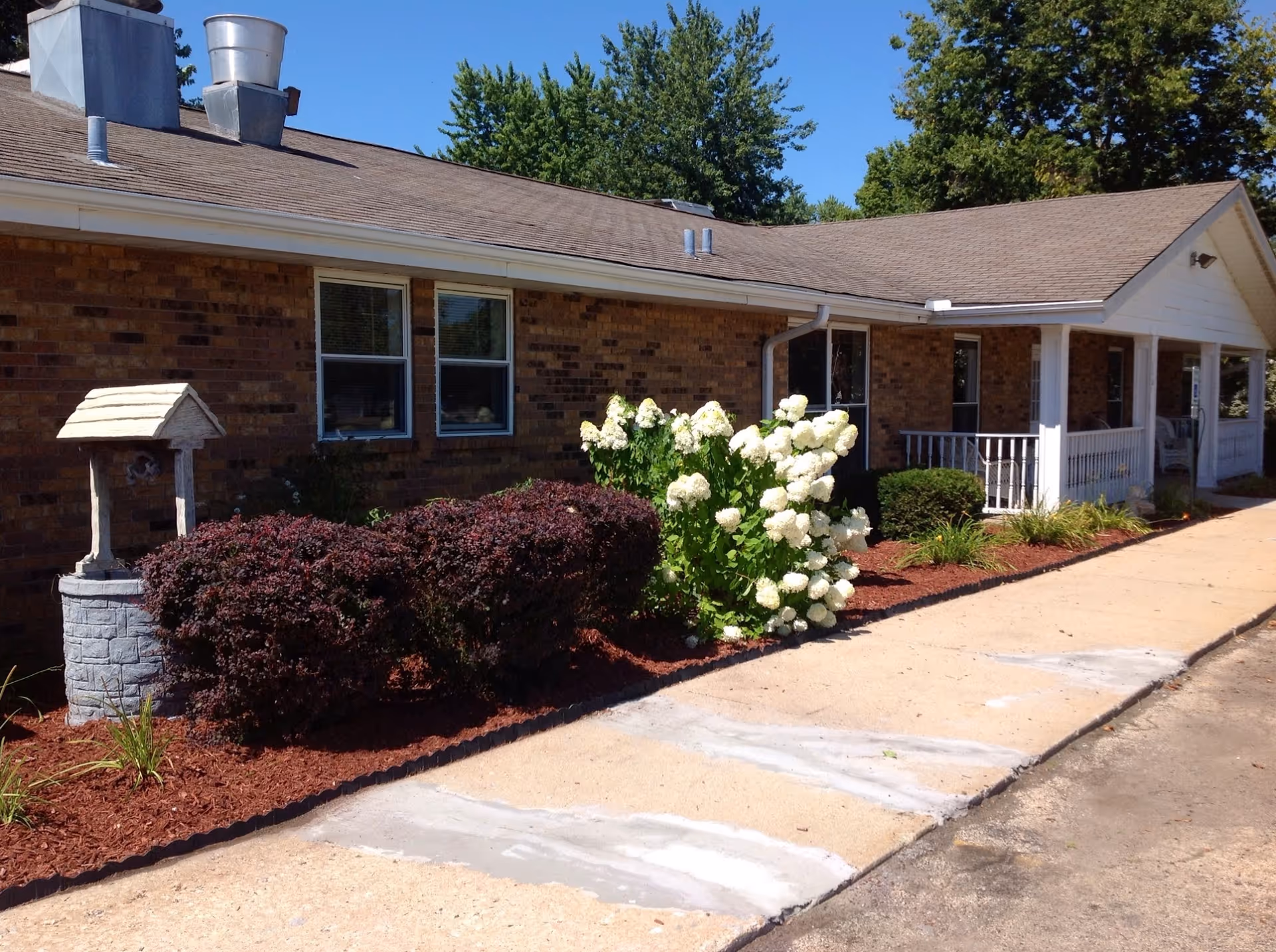 Front exterior of a single-story brick rehabilitation building with landscaped shrubs, white flowering bushes, and a covered porch by a sidewalk.
