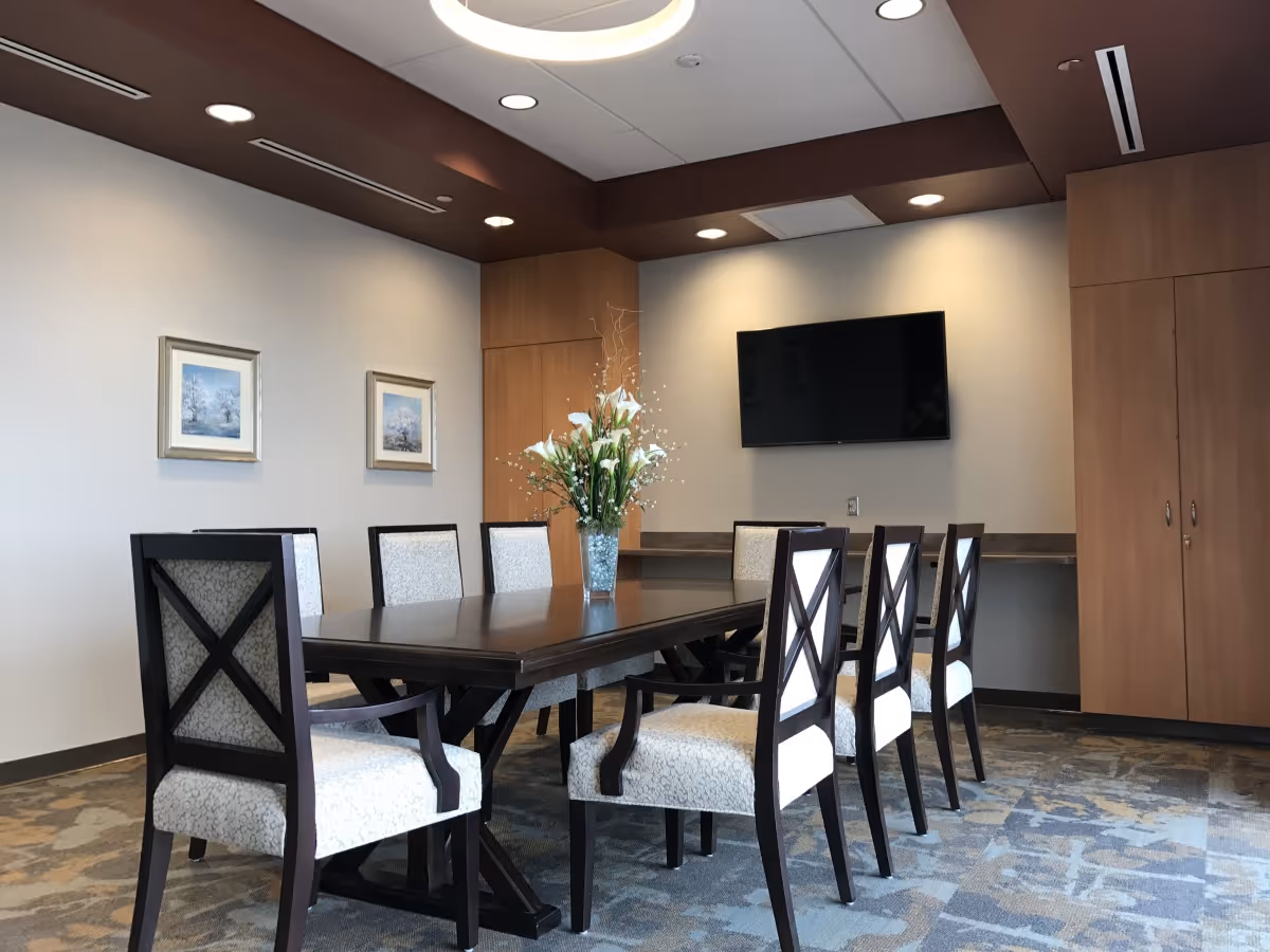 A modern dining room with a long dark wooden table surrounded by eight upholstered chairs with patterned fabric. A vase with white flowers is placed in the center of the table. The room has light-colored walls with two framed pictures, a mounted flat-screen TV, recessed lighting in the ceiling, and wooden cabinets along one wall.