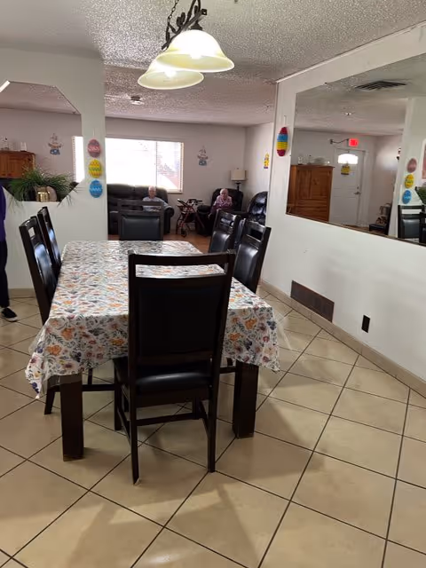 Interior view of a senior living facility dining area with a rectangular table covered with a floral tablecloth and surrounded by dark wooden chairs. In the background, there is a seating area with two elderly individuals sitting on recliners near a window. The room has tiled floors, white walls, and a large mirror on one side reflecting part of the room. Ceiling lights hang above the dining table.