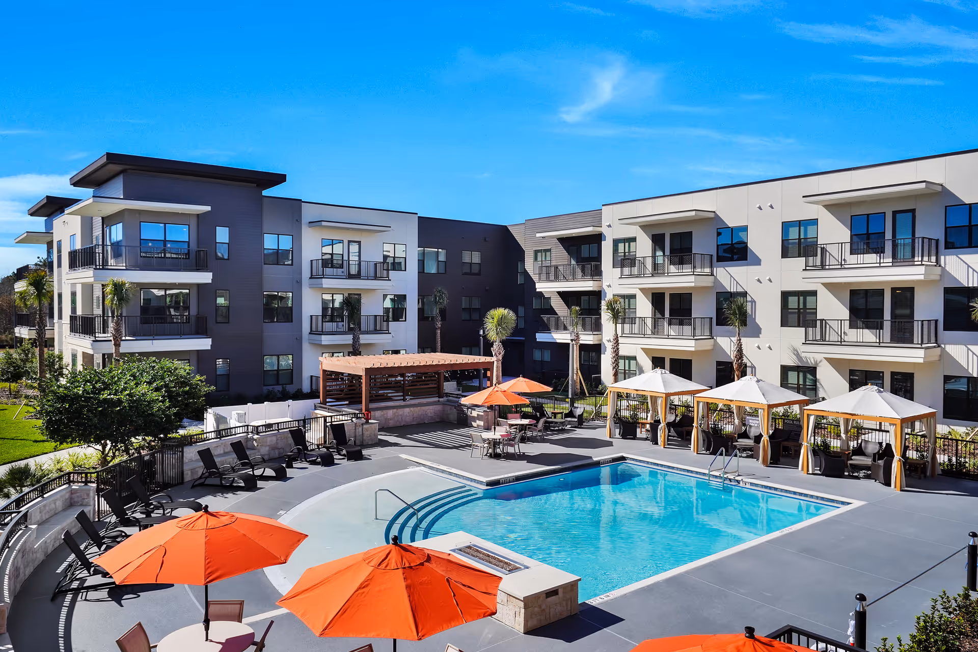Outdoor swimming pool area at Discovery Village Deerwood Independent Living with lounge chairs, orange umbrellas, cabanas, and a modern three-story building in the background under a clear blue sky.