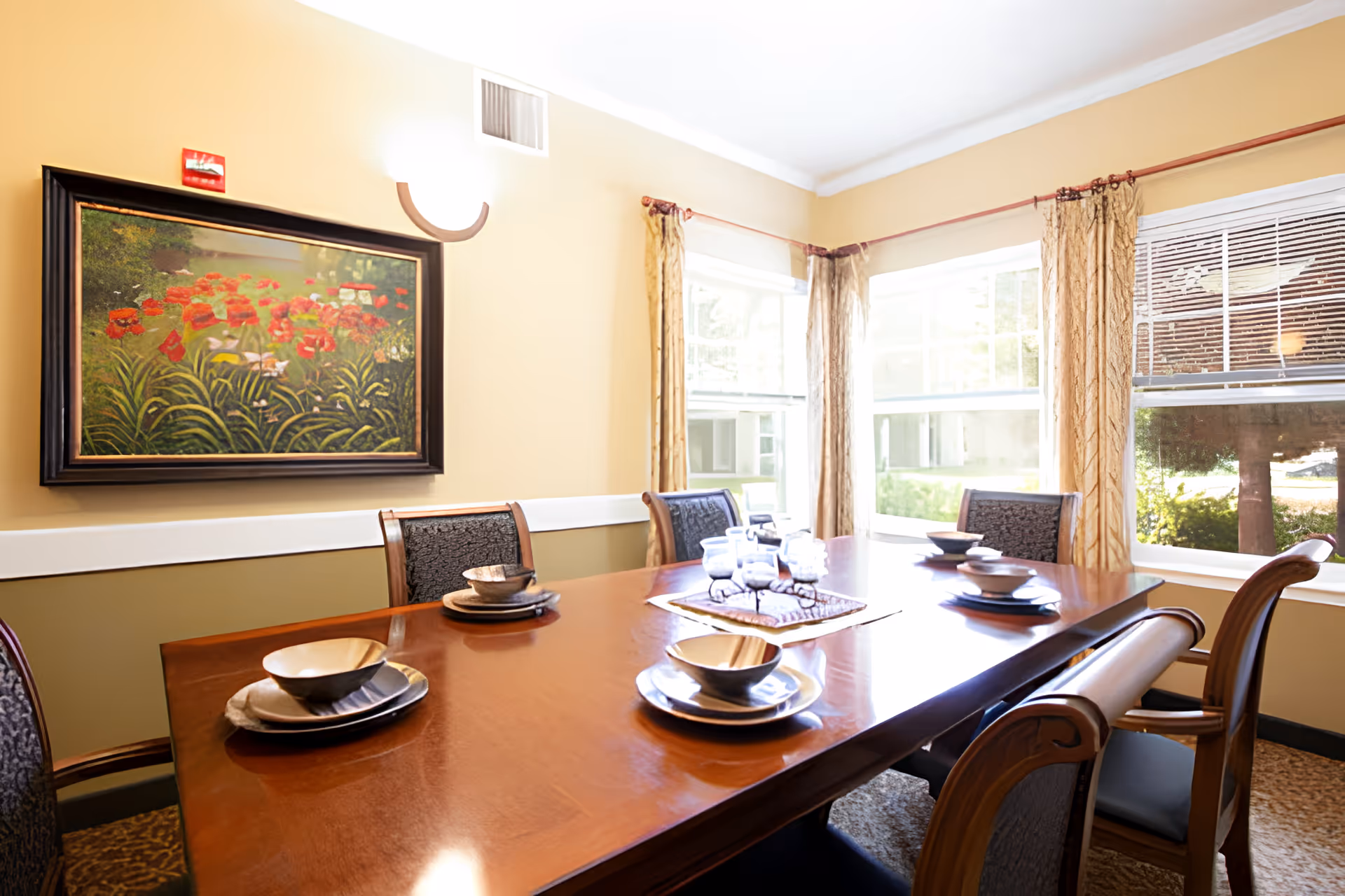 A dining room with a polished wooden table set with bowls and plates for six people. The room has large windows with beige curtains allowing natural light to fill the space. A framed painting of red flowers hangs on the beige wall.