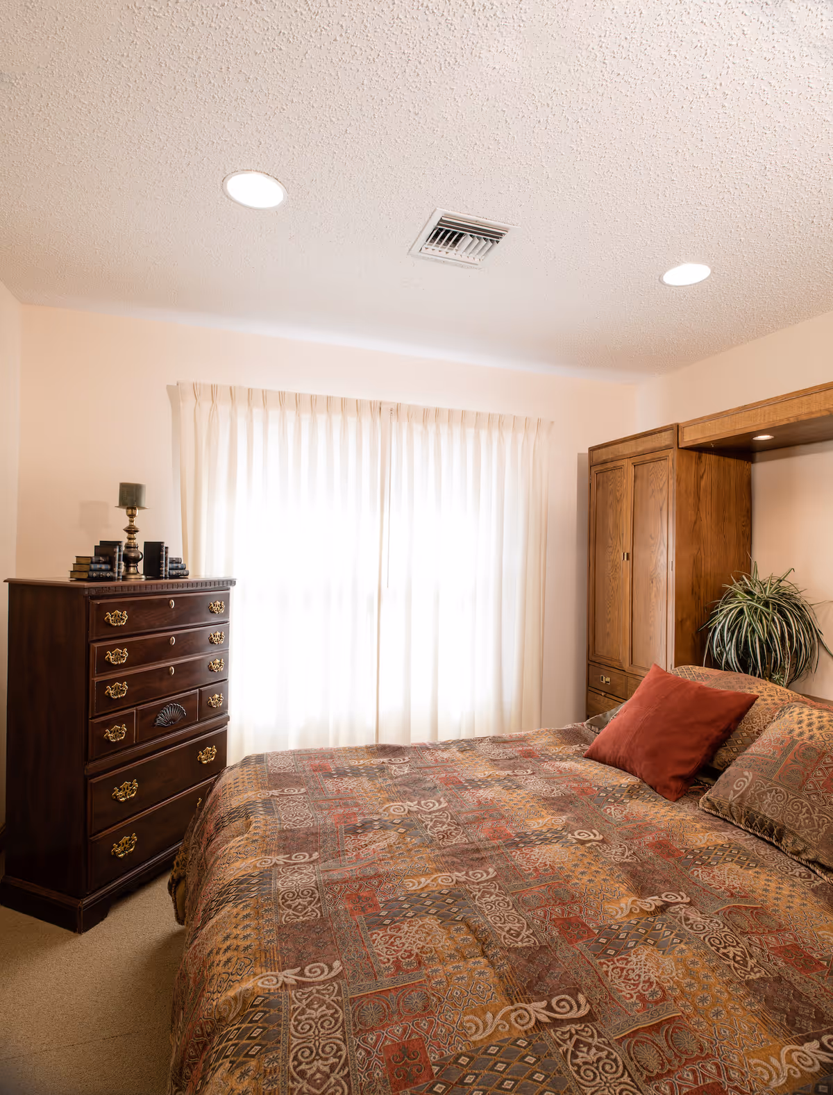 A cozy bedroom with a large bed covered in a patterned quilt and multiple pillows. To the left of the bed is a dark wooden chest of drawers with decorative handles, topped with a candle holder and some books. Behind the bed is a wooden headboard with built-in cabinets and a potted plant. A window with sheer white curtains allows natural light to softly illuminate the room.