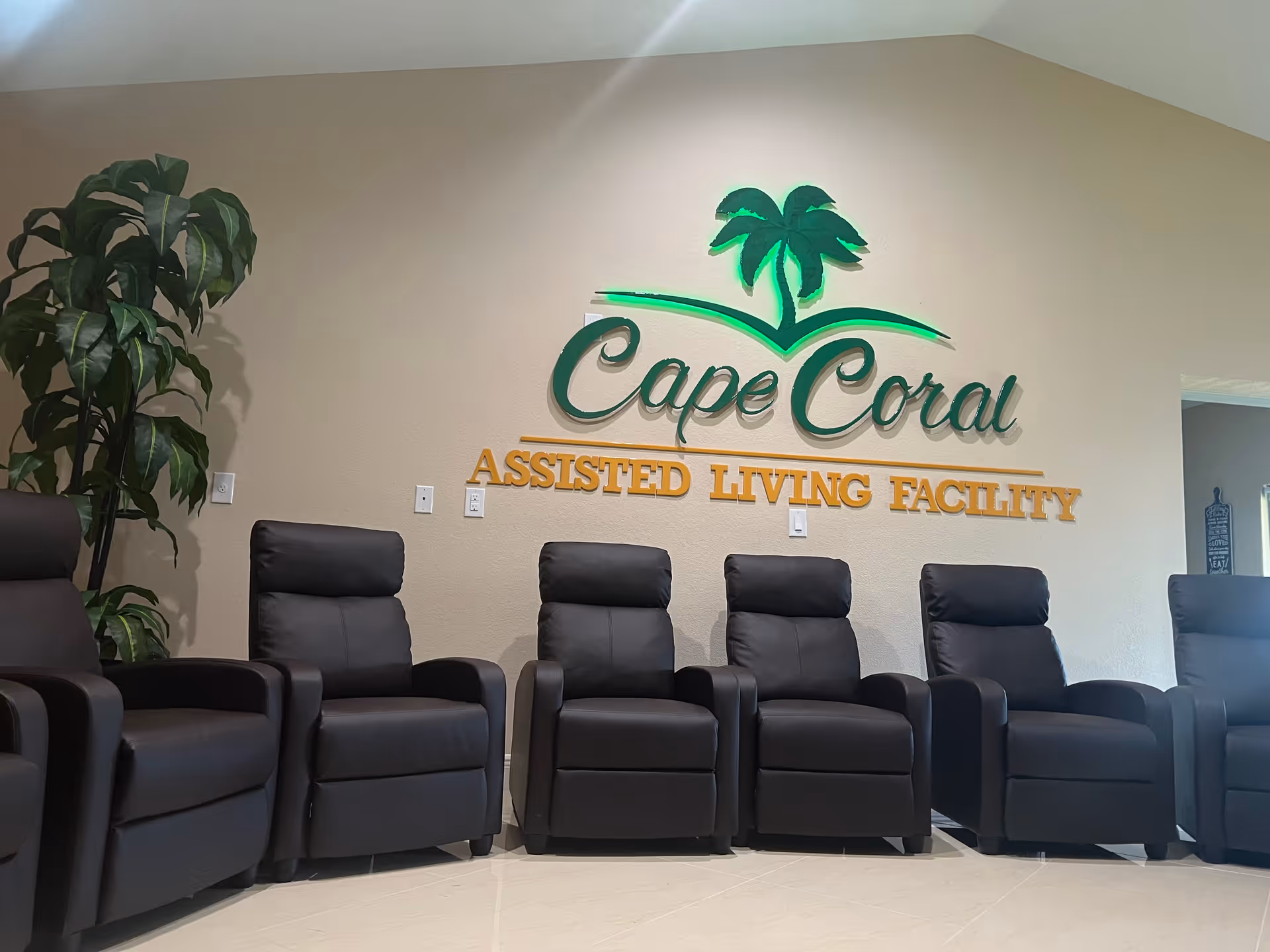 A row of six black recliner chairs lined up against a beige wall with a large green and yellow sign that reads 'Cape Coral Assisted Living Facility' and features a green palm tree logo. There is a tall green plant on the left side of the chairs.