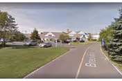 Driveway leading to the front facade of a low-rise residential building with pitched roofs, parked cars, and lawns.