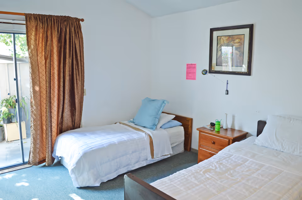 Bright shared bedroom with two single beds, a wooden nightstand, a sliding glass door with brown curtains, and framed art on the wall.