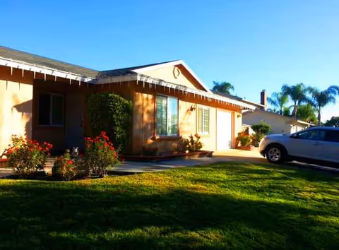 A single-story beige facility building with a manicured lawn and a parked car in the driveway.