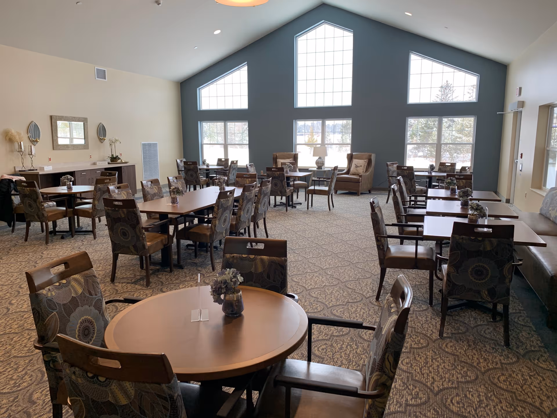A spacious dining room with multiple wooden tables and cushioned chairs arranged neatly. Large windows with geometric shapes allow natural light to fill the room. There are decorative plants on the tables and a sideboard against the wall with a mirror and wall sconces above it. Two armchairs with pillows are placed near the windows.