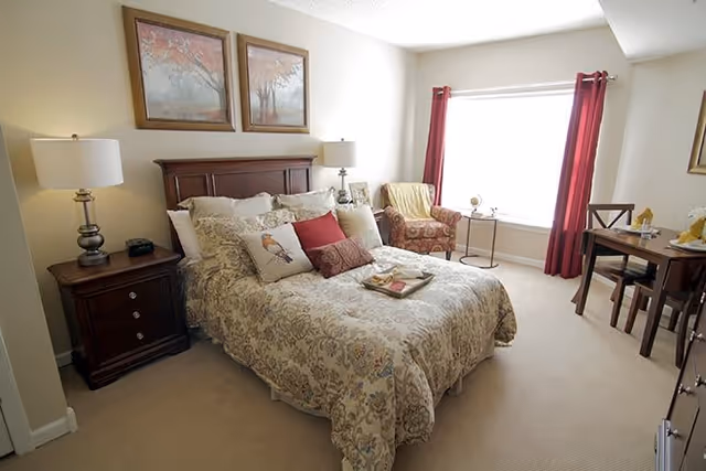A cozy bedroom with a large bed covered in a floral patterned comforter and multiple pillows. There are two framed paintings of trees above the headboard. On either side of the bed are wooden nightstands with lamps. A patterned armchair and a small round table are positioned near a large window with red curtains. A wooden dining table with chairs is visible on the right side of the room.
