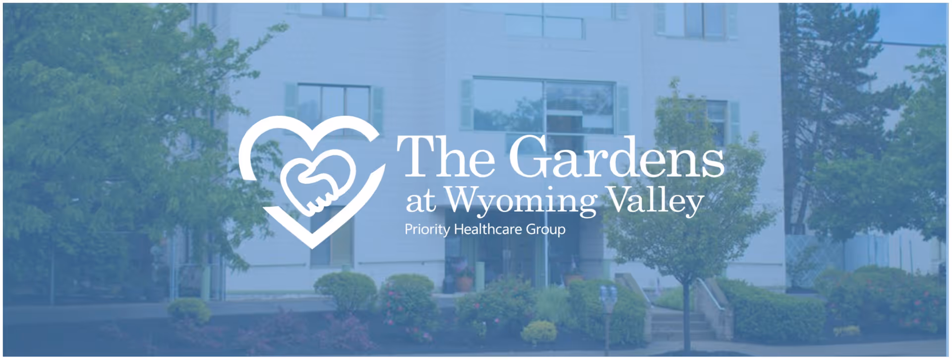 Exterior view of a multi-story building surrounded by trees and shrubs with a logo overlay showing two hands forming a heart and text that reads 'The Gardens at Wyoming Valley Priority Healthcare Group'.