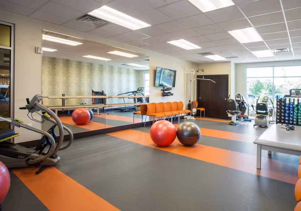 Bright exercise room with cardio machines, two exercise balls, mirrored wall, and orange seating.