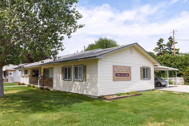 Single-story building with light-colored siding and a shingled roof, surrounded by green grass and trees. A sign on the side of the building reads Ashley Manor Memory Care with a phone number and website. There is a carport on the right side with a vehicle parked underneath.