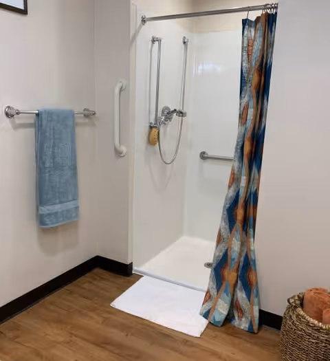 A clean and accessible shower area with a white shower base, grab bars, a handheld showerhead, and a colorful shower curtain. A blue towel hangs on a towel rack on the adjacent wall, and a white bath mat is placed in front of the shower. The floor is wood-style, and a woven basket with towels is visible in the corner.