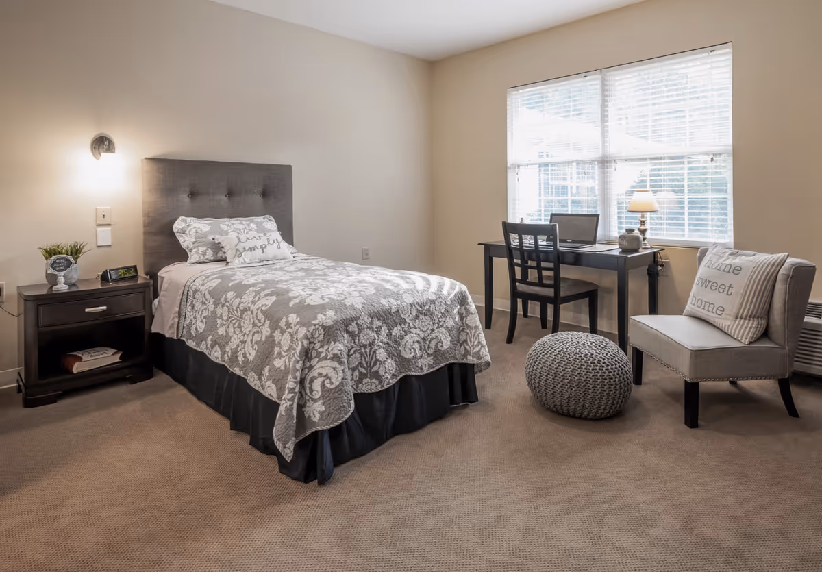 A neatly arranged bedroom with a single bed covered in a gray and white patterned quilt and pillows, a dark wooden nightstand with a small plant and digital clock, a black desk with a chair and a laptop near a window with white blinds, and a gray upholstered chair with a 'home sweet home' pillow next to a knitted pouf on a beige carpeted floor.