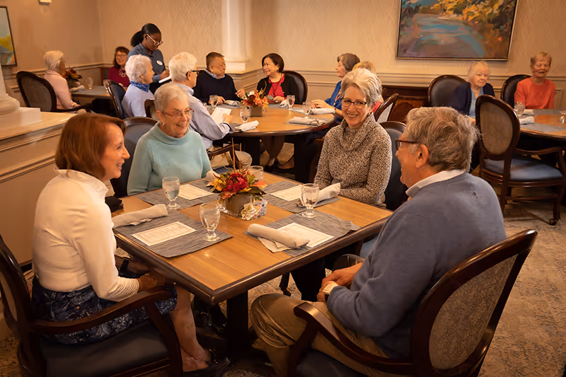 A group of elderly people sitting at tables in a warmly lit dining room, engaging in conversation and smiling. The tables are set with placemats, napkins, glasses, and a small floral centerpiece. The room has beige walls, framed artwork, and a carpeted floor.