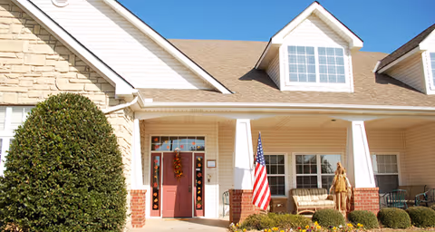 Front exterior view of a senior living facility with a beige and brick facade, a red front door decorated with autumn wreaths, an American flag displayed near the entrance, and a porch with seating and landscaping including bushes and flowers.