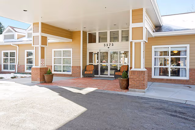 Entrance of a senior living facility with a covered drop-off area, two chairs, potted plants, and large windows on either side of double glass doors. The building exterior features beige siding with brick accents and the number 3375 above the entrance.