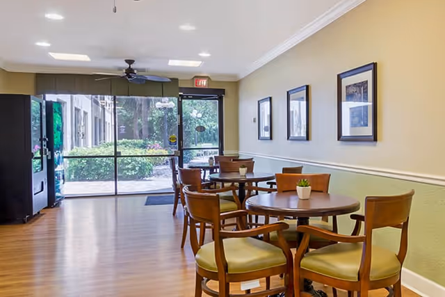 A bright common area with wooden round tables and chairs, small potted plants on the tables, framed pictures on the wall, a ceiling fan, and large glass doors leading outside with greenery visible.
