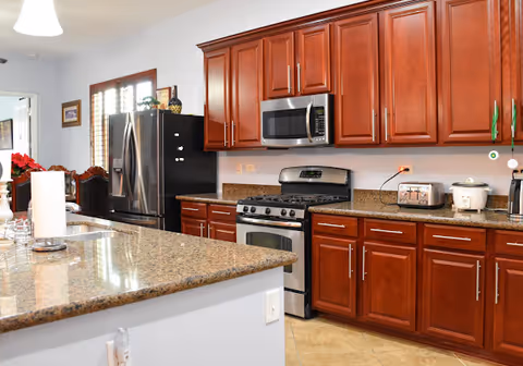 A modern kitchen with wooden cabinets, granite countertops, a stainless steel refrigerator, stove, microwave, toaster, and rice cooker. The kitchen has tiled floors and a window with wooden shutters.