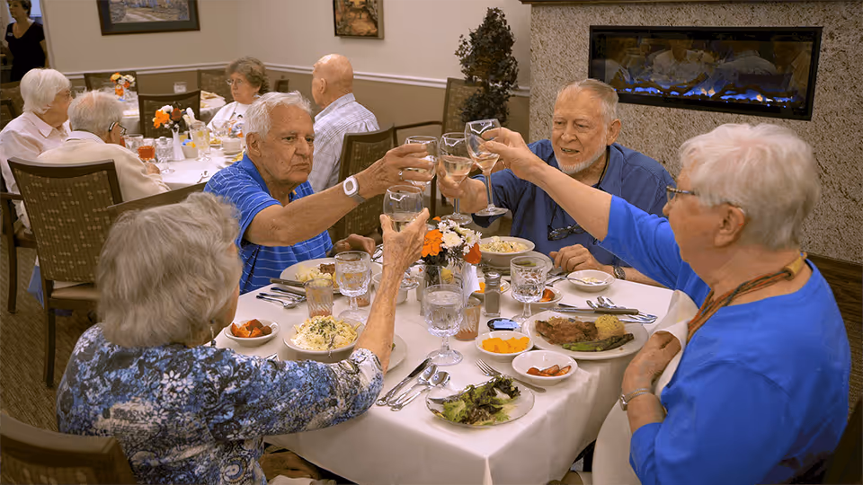 A group of elderly people sitting around a dining table in a senior living facility, raising their glasses in a toast. The table is set with plates of food, bowls of salad and fruit, and glasses of water. There is a fireplace on the wall behind them and other residents dining in the background.