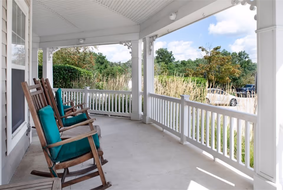 A covered porch area with white railings and columns, featuring three wooden rocking chairs with green cushions. The porch overlooks a landscaped area with tall grasses and trees, and a parking lot with cars is visible in the background under a partly cloudy sky.