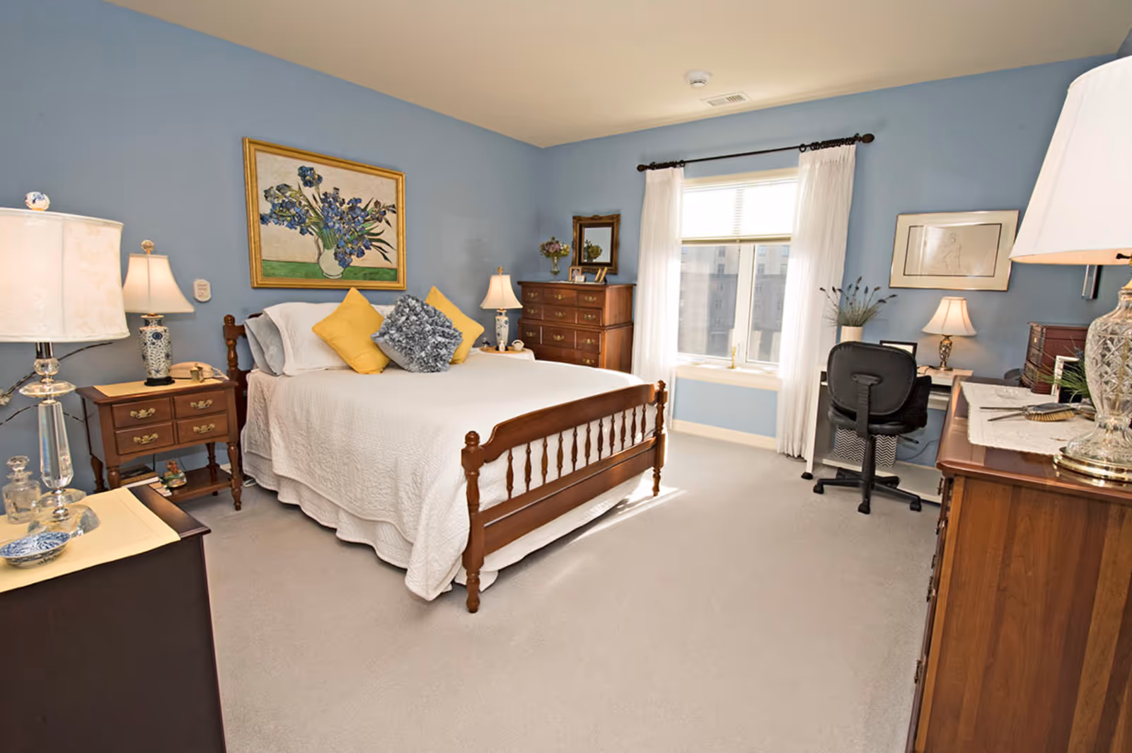 A cozy bedroom with light blue walls, a wooden bed with white bedding and yellow and gray pillows, two wooden nightstands with lamps, a wooden dresser, a window with white curtains, a desk with a black office chair, and framed artwork on the walls.