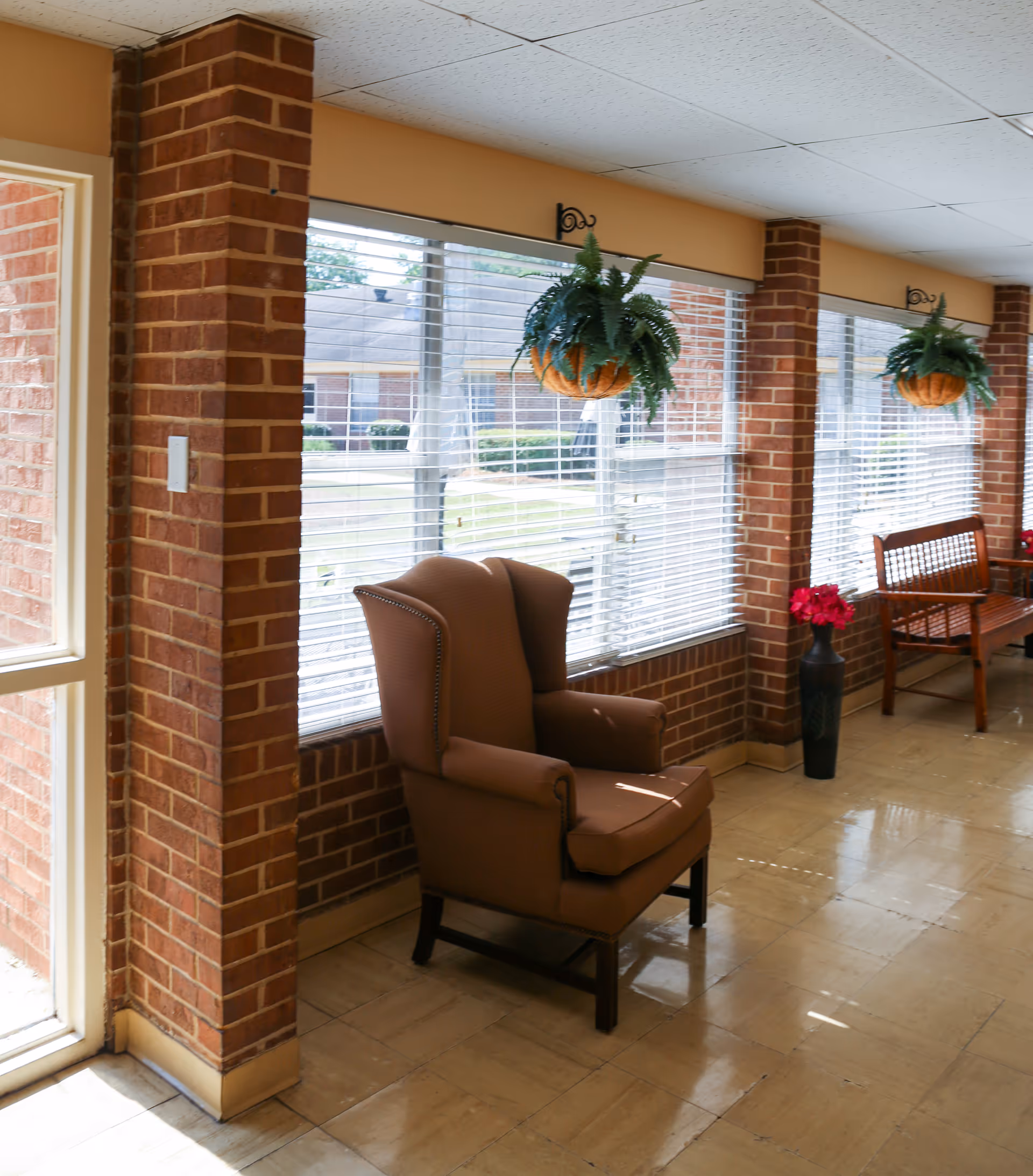 A bright indoor seating area with large windows covered by white blinds. There is a brown upholstered armchair and a wooden bench along the brick wall. Two hanging baskets with green plants and a tall vase with red flowers are also visible.