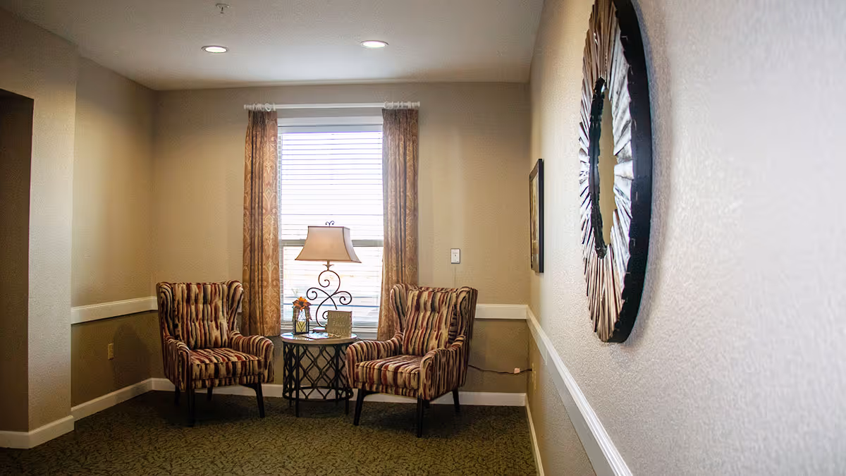 A cozy sitting area in a senior living facility with two patterned armchairs facing each other, a small round table between them holding a decorative lamp, a small flower arrangement, and a sign. The area is lit by natural light coming through a window with curtains. A large decorative circular mirror hangs on the adjacent wall.