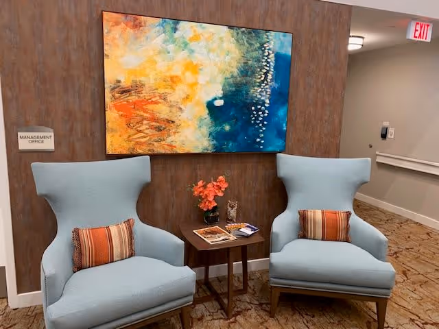 Two light blue upholstered armchairs with striped orange and brown pillows are positioned on either side of a small wooden table holding a vase with orange flowers and some magazines. Behind the chairs is a brown wall with a colorful abstract painting featuring shades of yellow, orange, blue, and white. A sign on the wall reads 'Management Office'. The floor is carpeted with a patterned design, and a hallway with an exit sign is visible to the right.