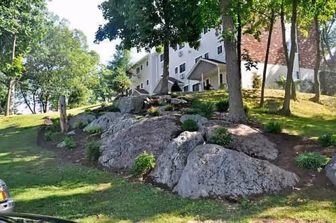 A grassy outdoor area with large rocks and several trees in front of a multi-story white building with a brick section and multiple windows.