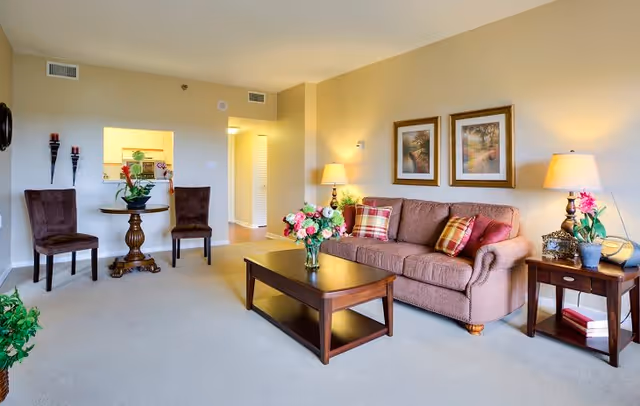 A cozy living room with a brown sofa adorned with plaid and solid pillows, a wooden coffee table with a vase of flowers, two framed pictures on the wall, a side table with a lamp and decorative items, and two chairs with a small round table near a pass-through window to the kitchen.