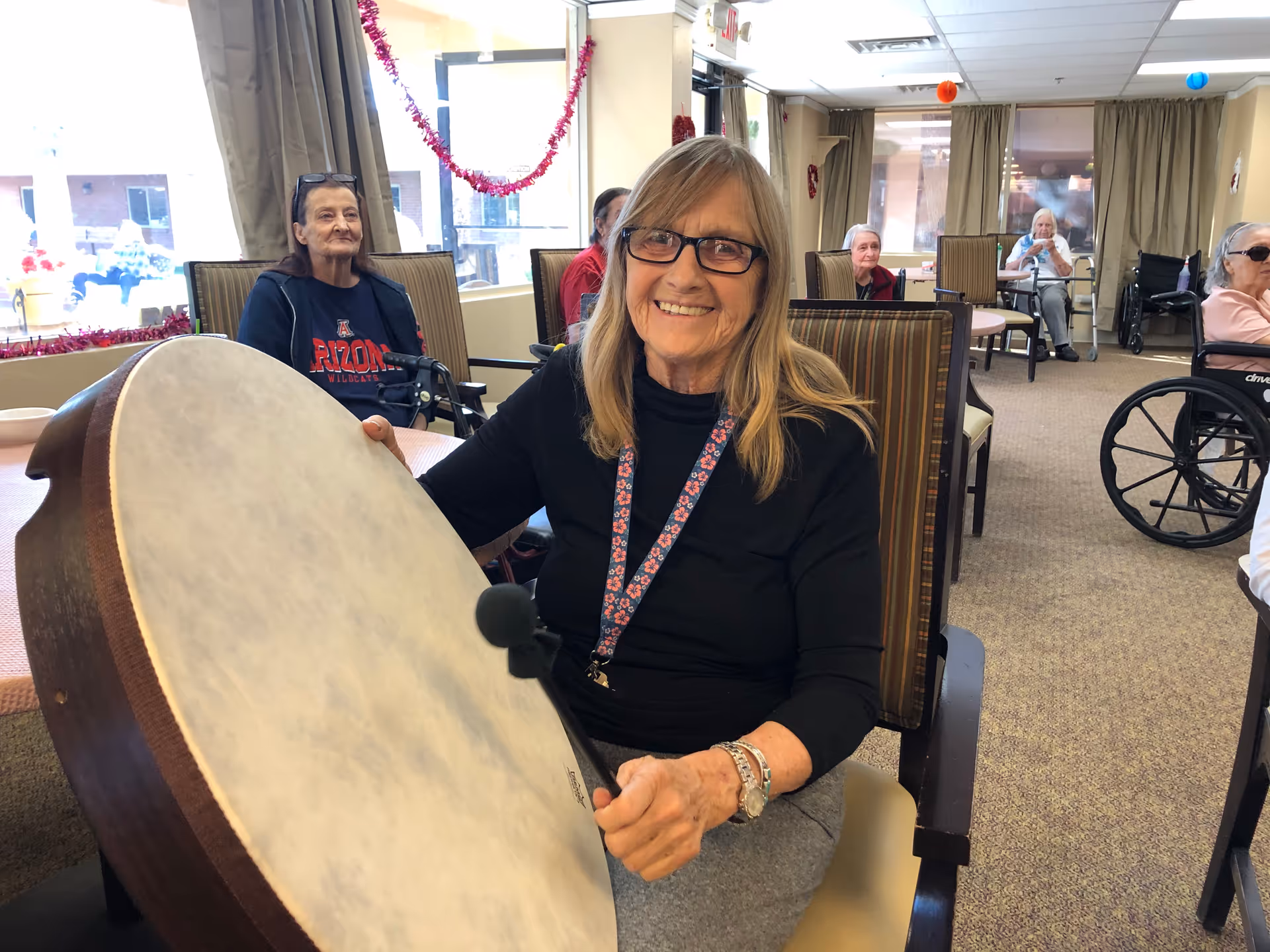 An elderly woman smiling and holding a large drum with a mallet in a communal room. Other elderly individuals are seated around tables in the background, some in wheelchairs. The room has large windows with curtains and festive decorations hanging from the ceiling and walls.