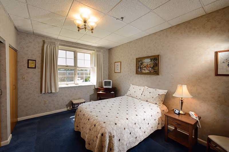 A cozy bedroom with a double bed covered in a white quilt with a geometric pattern. The room has beige wallpaper, a blue carpet, and a window with cream curtains letting in natural light. There is a wooden nightstand with a lamp, a telephone, and a clock beside the bed. A small TV sits on a wooden stand in the corner near the window. Framed artwork hangs on the walls.