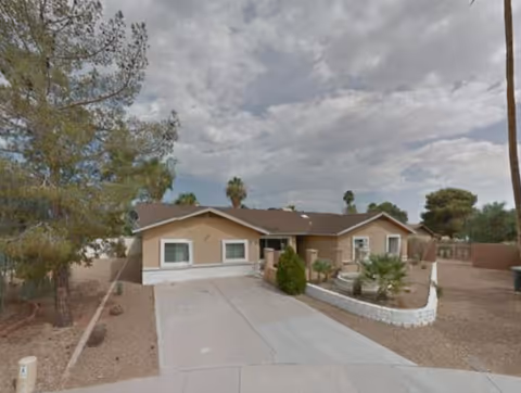 Single-story residential building with a beige exterior and brown roof, surrounded by desert landscaping including trees, bushes, and gravel. The driveway leads up to the entrance, and the sky is partly cloudy.