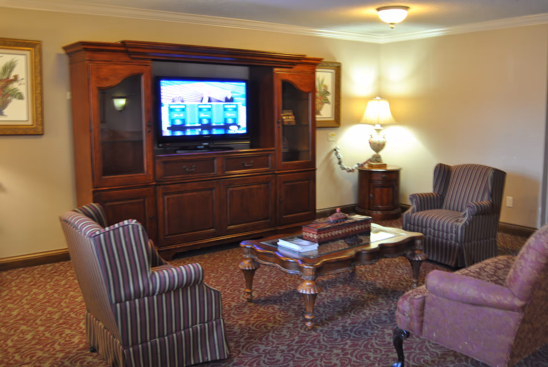 A cozy living room with a wooden entertainment center holding a flat-screen TV, surrounded by three upholstered armchairs with striped and patterned fabric. A wooden coffee table with books and a decorative box sits in the center on a patterned carpet. A side table with a lamp and framed botanical prints adorn the beige walls.