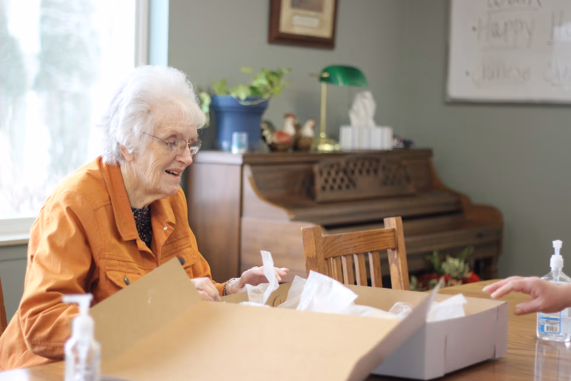 An elderly woman with white hair and glasses, wearing an orange jacket, is sitting at a wooden table and smiling while unpacking a large cardboard box. In the background, there is a wooden piano with decorative items on top, a green desk lamp, and a potted plant. A window is visible to the left, and a whiteboard with some writing is on the wall behind her. A bottle of hand sanitizer is on the table.