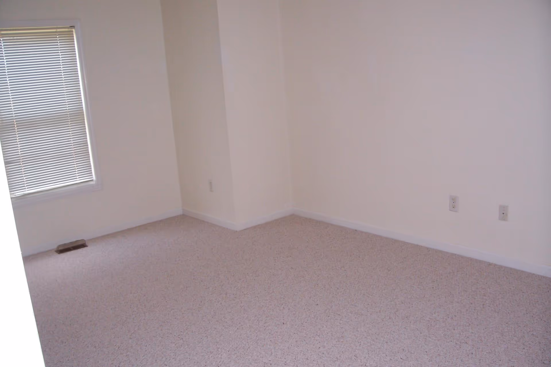 Empty carpeted room with beige walls, a window with closed blinds, and electrical outlets along the wall.