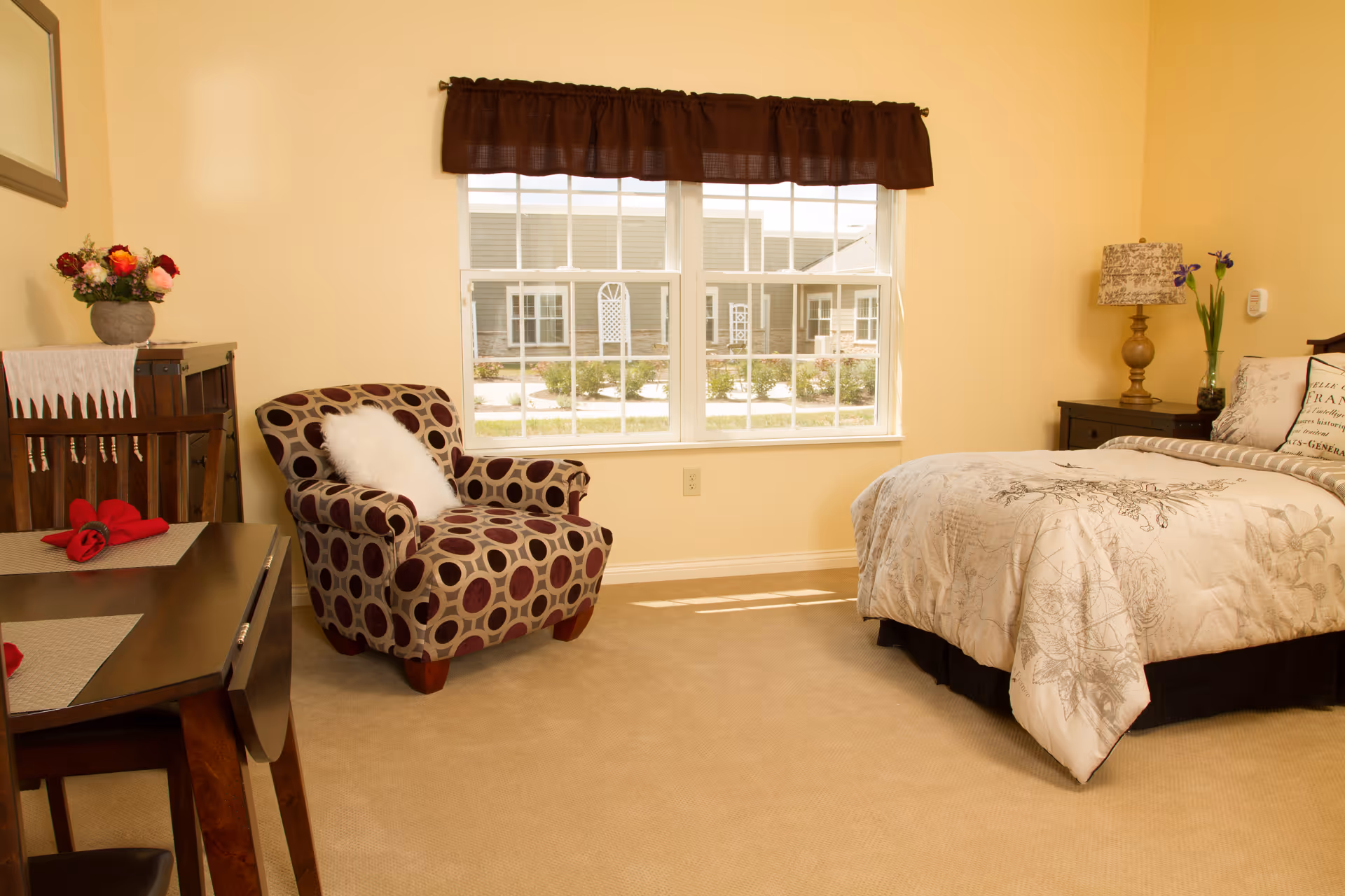 Sunlit bedroom with a bed, patterned armchair, small table with flowers, and a window overlooking a courtyard.